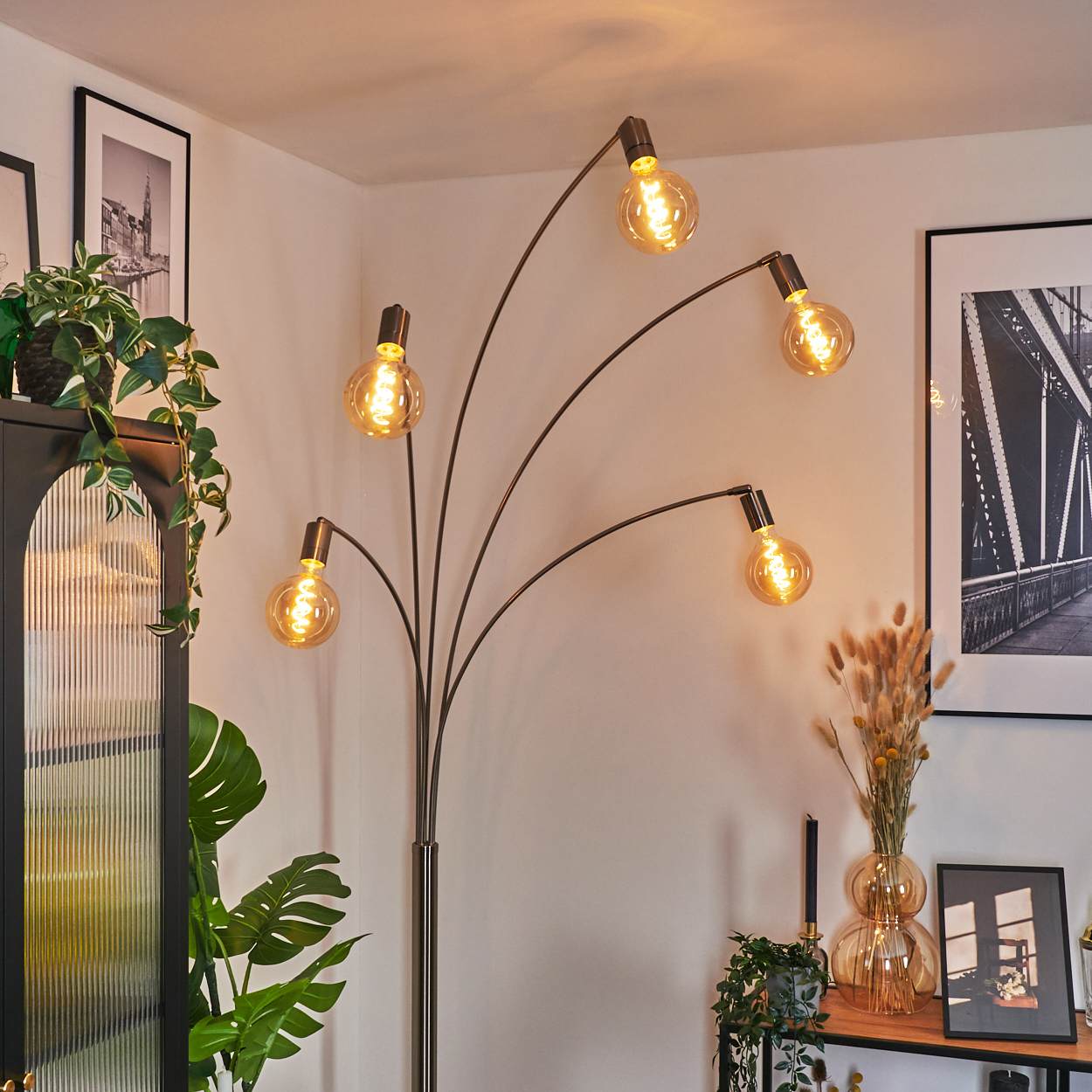 Eine moderne Stehlampe mit fünf leuchtenden Glühbirnen. Sie steht in einem Raum mit Pflanzen, Bildern und einem Holztisch. Die Wand ist weiß.