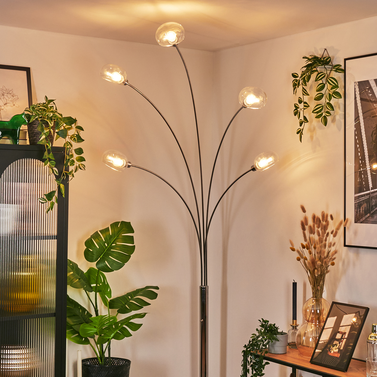 Ein modernes Wohnzimmer mit Stehlampe, Pflanzen und gerahmter Kunst. Die Lampe hat fünf Glühbirnen. Pflanzen in Töpfen und hängen.