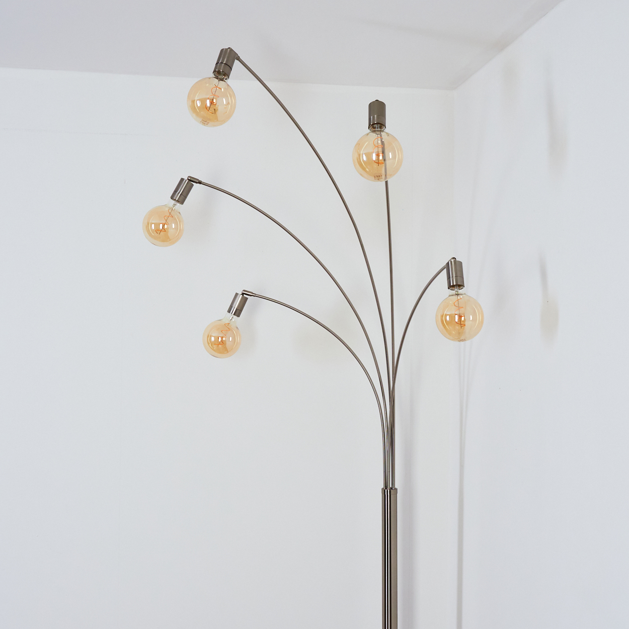 Eine moderne Stehlampe mit fünf runden, leuchtenden Glühbirnen. Die Lampe hat ein silbernes Gestell vor einer weißen Wand.