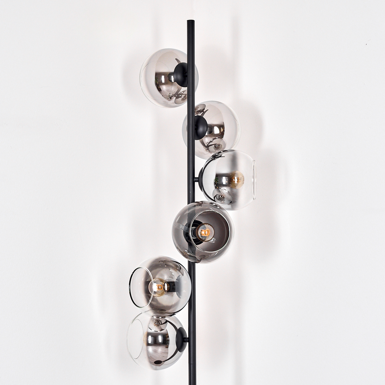 Eine moderne Wandlampe mit sechs Glaskugelschirmen in verschiedenen Grautönen, befestigt an einer schwarzen Metallsäule, vor einer weißen Wand.