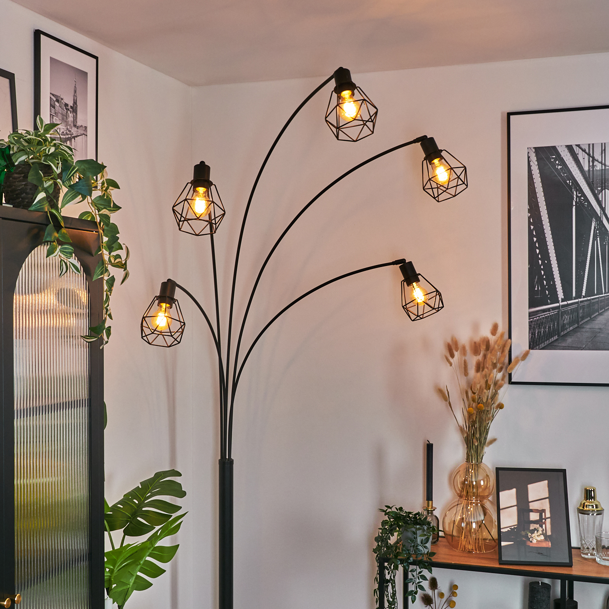 Eine schwarze Stehlampe mit fünf Lichtern. Der Hintergrund umfasst gerahmte Kunst, Pflanzen und ein Regal mit Dekor.