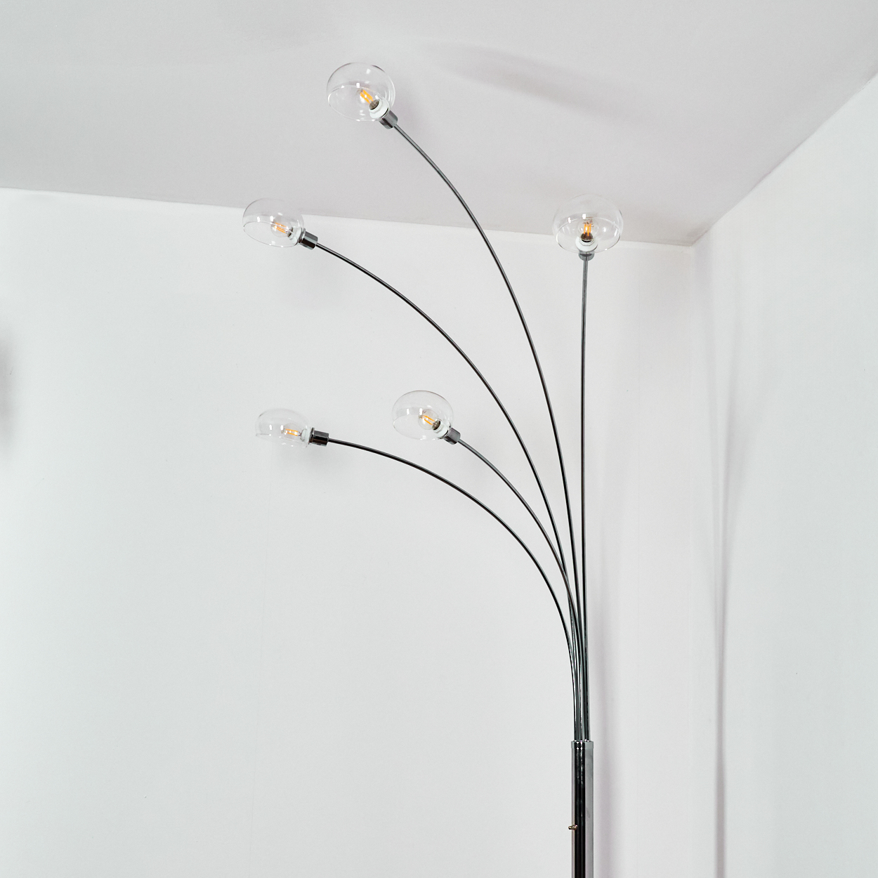 Eine moderne Stehlampe mit fünf klaren Glaskugelschirmen. Die Lampe hat ein Chromgestell und schwarze, gebogene Arme vor einer weißen Wand.