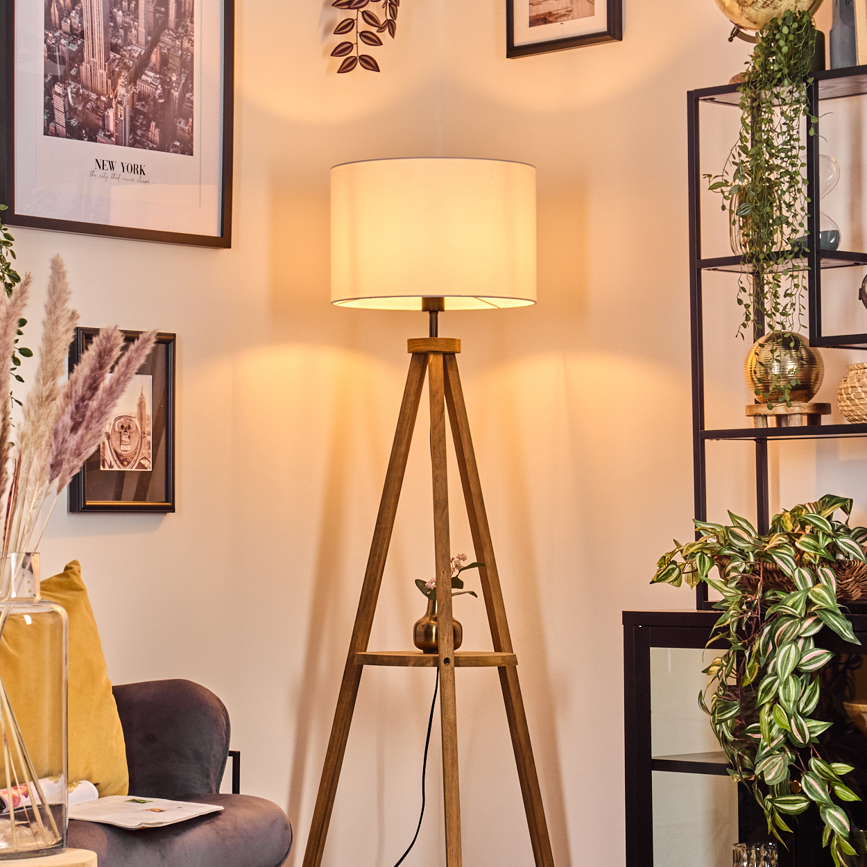 Holzstativlampe mit weißem Lampenschirm. Ein Regal hält eine Vase. Dahinter sind Bilder, Pflanzen und ein Regal.