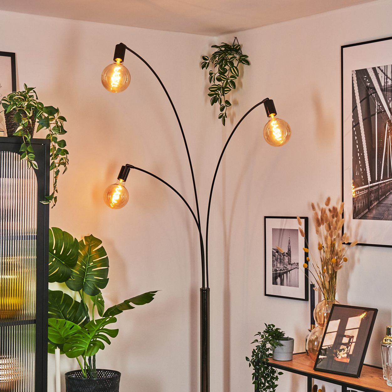 Eine moderne Stehlampe mit drei Glühbirnen ist eingeschaltet. Grüne Pflanzen sind in der Nähe. Bilder und ein Regal mit Gegenständen sind im Hintergrund.