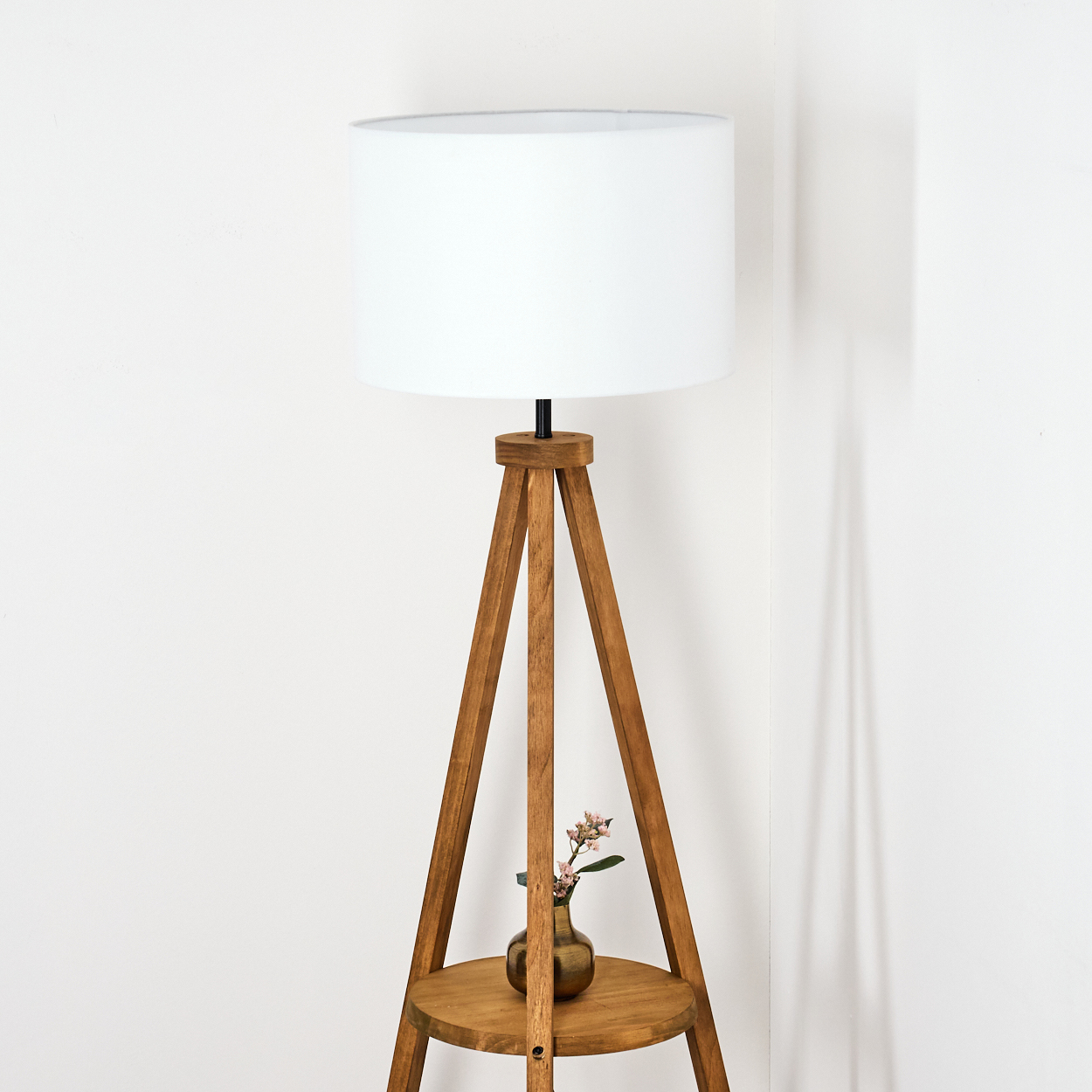 Eine hölzerne Stehlampe mit weißem Lampenschirm. Eine kleine Vase mit Blumen steht auf einem Regal. Der Hintergrund ist eine weiße Wand.