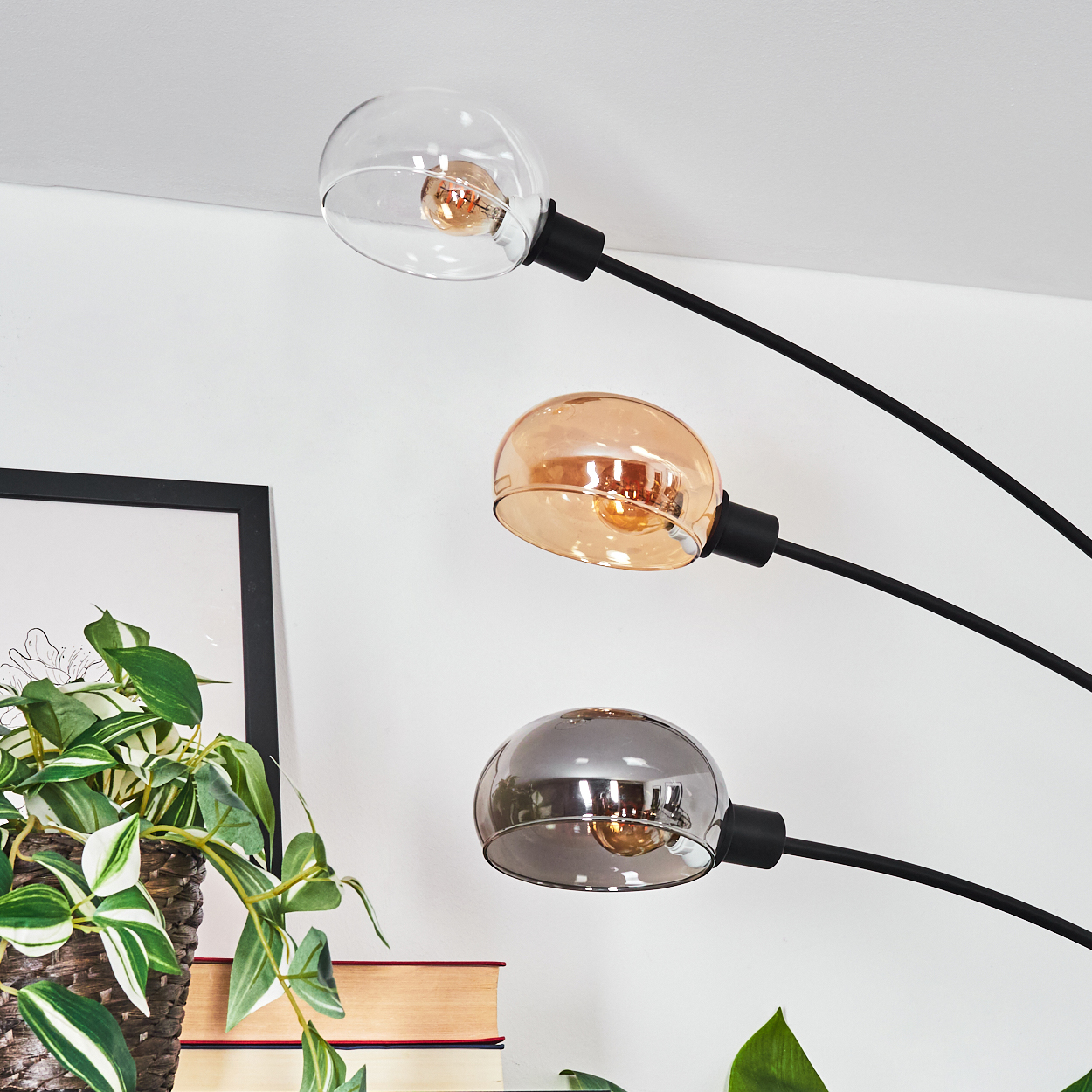 Lampe mit drei Glühbirnen mit transparenten, bernsteinfarbenen und silbernen Schirmen. Eine Zimmerpflanze und Bücher sind sichtbar.