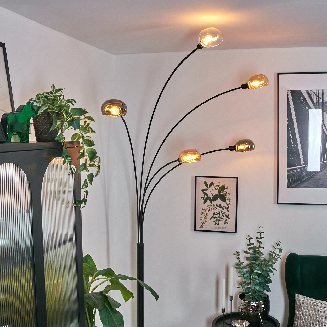 Ein modernes Wohnzimmer mit einer schwarzen Stehlampe mit fünf Glühbirnen. Pflanzen und Kunst sind ebenfalls sichtbar. Rechts ist ein grüner Sessel.