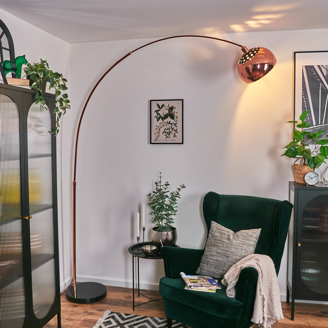 Modernes Wohnzimmer mit grünem Sessel, Kupferlampe und Pflanzen. Kunstwerk und schwarzer Schrank sind sichtbar.