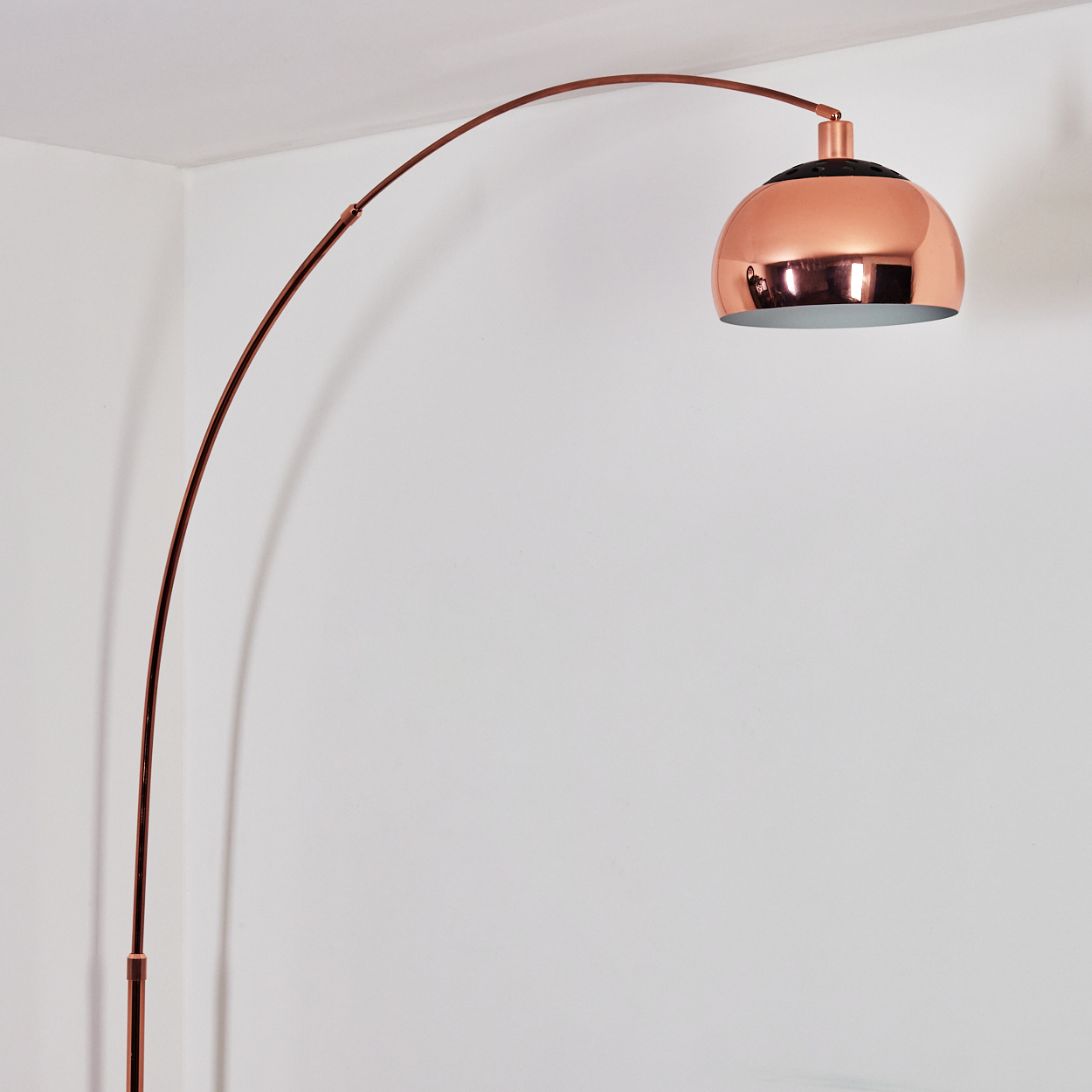 Eine kupferfarbene Bogenstehlampe vor einer weißen Wand. Die Lampe hat einen großen, runden Schirm und einen gebogenen Arm.