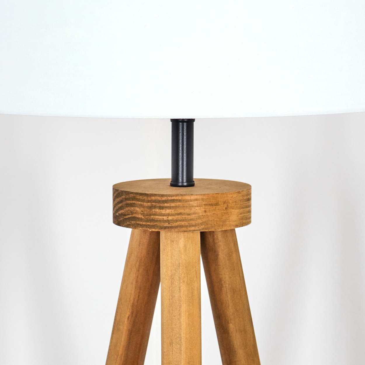 Eine Holzlampe mit weißem Lampenschirm. Sie hat einen schwarzen Metallstiel und ein Stativ. Der Hintergrund ist eine weiße Wand.