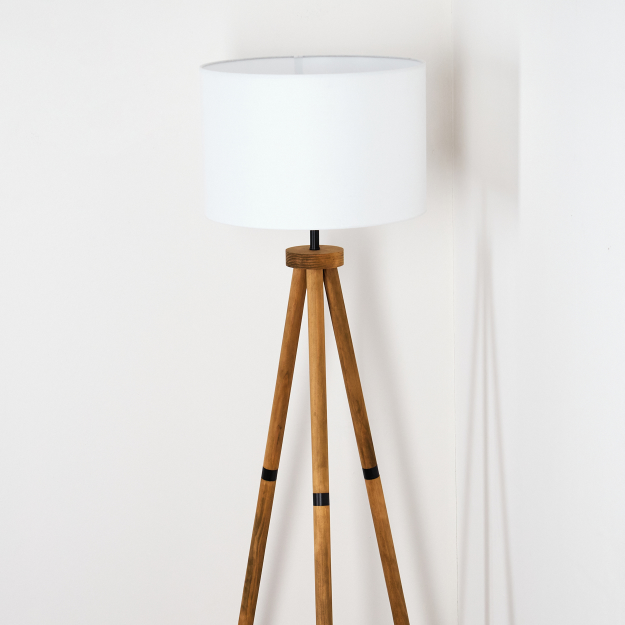 Eine Stehlampe mit Holzstativ und weißem Lampenschirm steht vor einer weißen Wand. Die Lampe hat einen zylindrischen Schirm und Holzbeine.