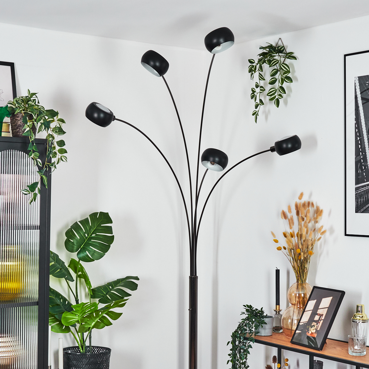 Ein modernes Wohnzimmer mit schwarzer Stehlampe, Pflanzen, Regal mit Dekorationen und einem schwarz-weißen Bild.