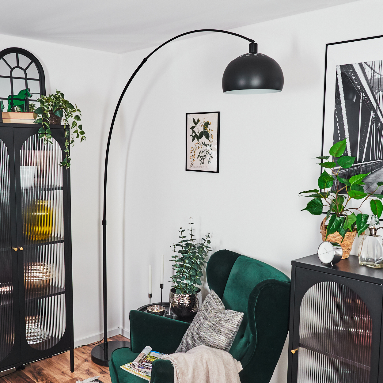 Ein Raum mit grünem Samtsessel, schwarzen Schränken, einer großen schwarzen Lampe und verschiedenen Pflanzen. Ein Brückenbild hängt an der Wand.