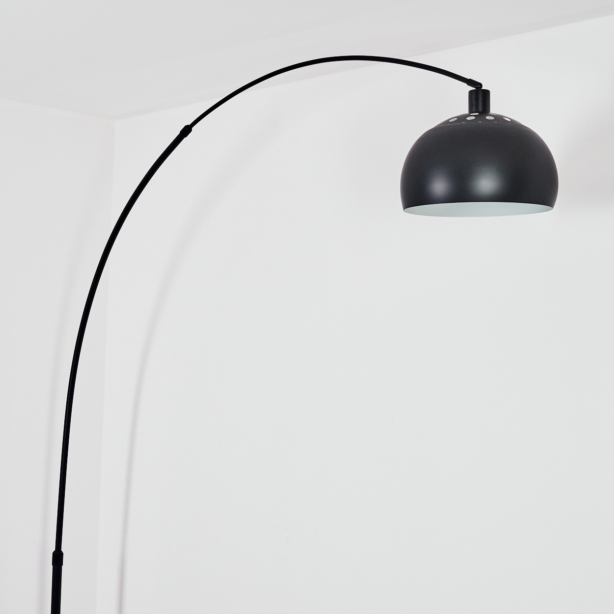 Eine schwarze Bogenstehlampe. Die Lampe hat einen kuppelförmigen Kopf, der vor einer weißen Wand in einer Ecke des Raumes positioniert ist.