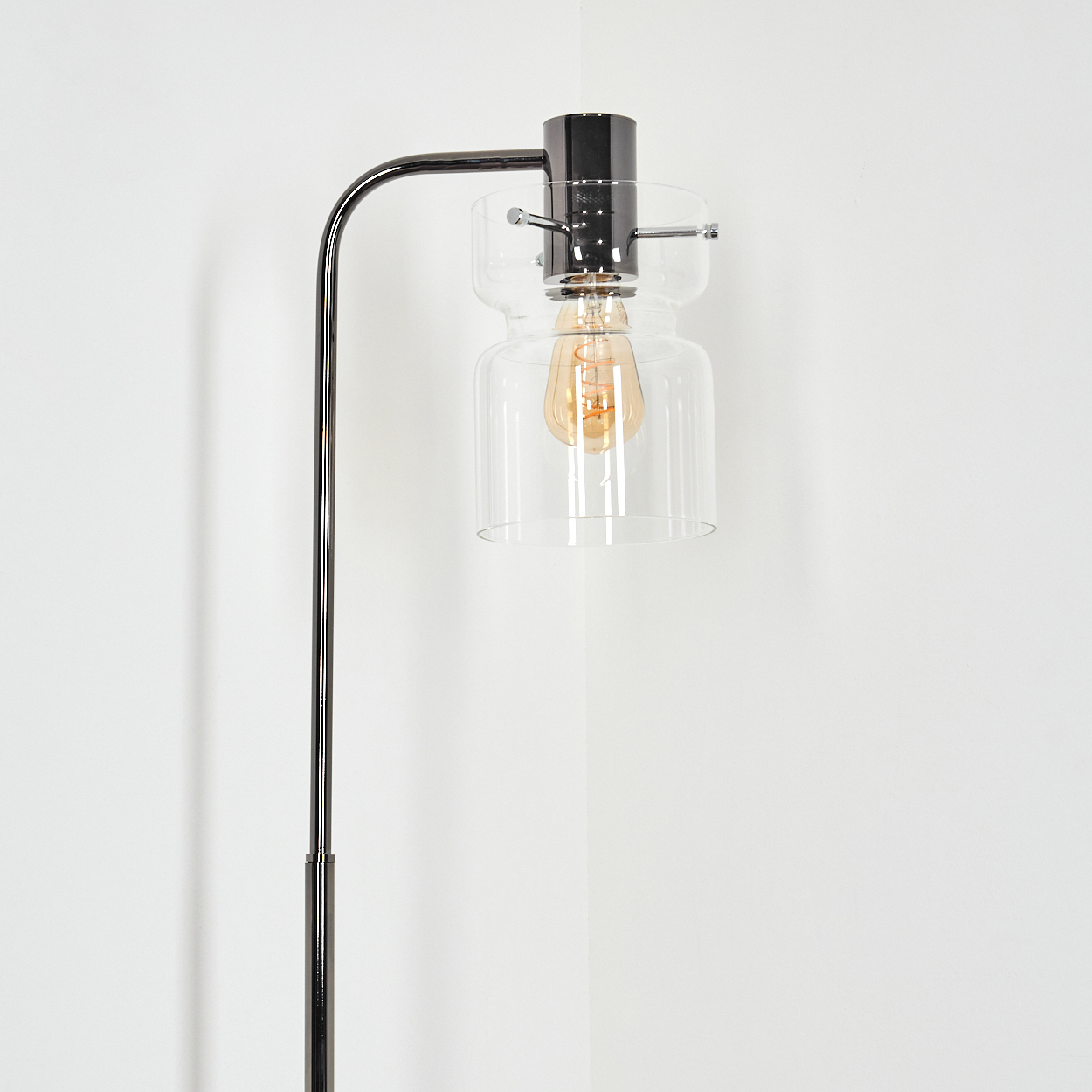 Eine moderne Stehlampe mit schwarzem Metallrahmen, Glasschirm und sichtbarer Glühbirne vor einer weißen Wand.
