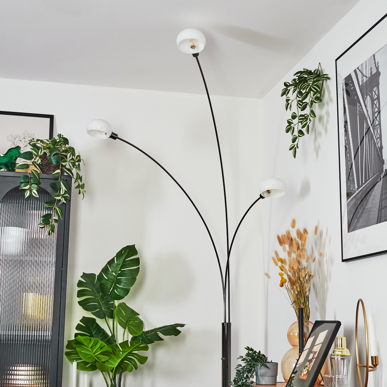 Ein modernes Wohnzimmer mit einer schwarzen Stehlampe mit drei weißen Lampenschirmen. Pflanzen und gerahmte Bilder schmücken die weißen Wände.