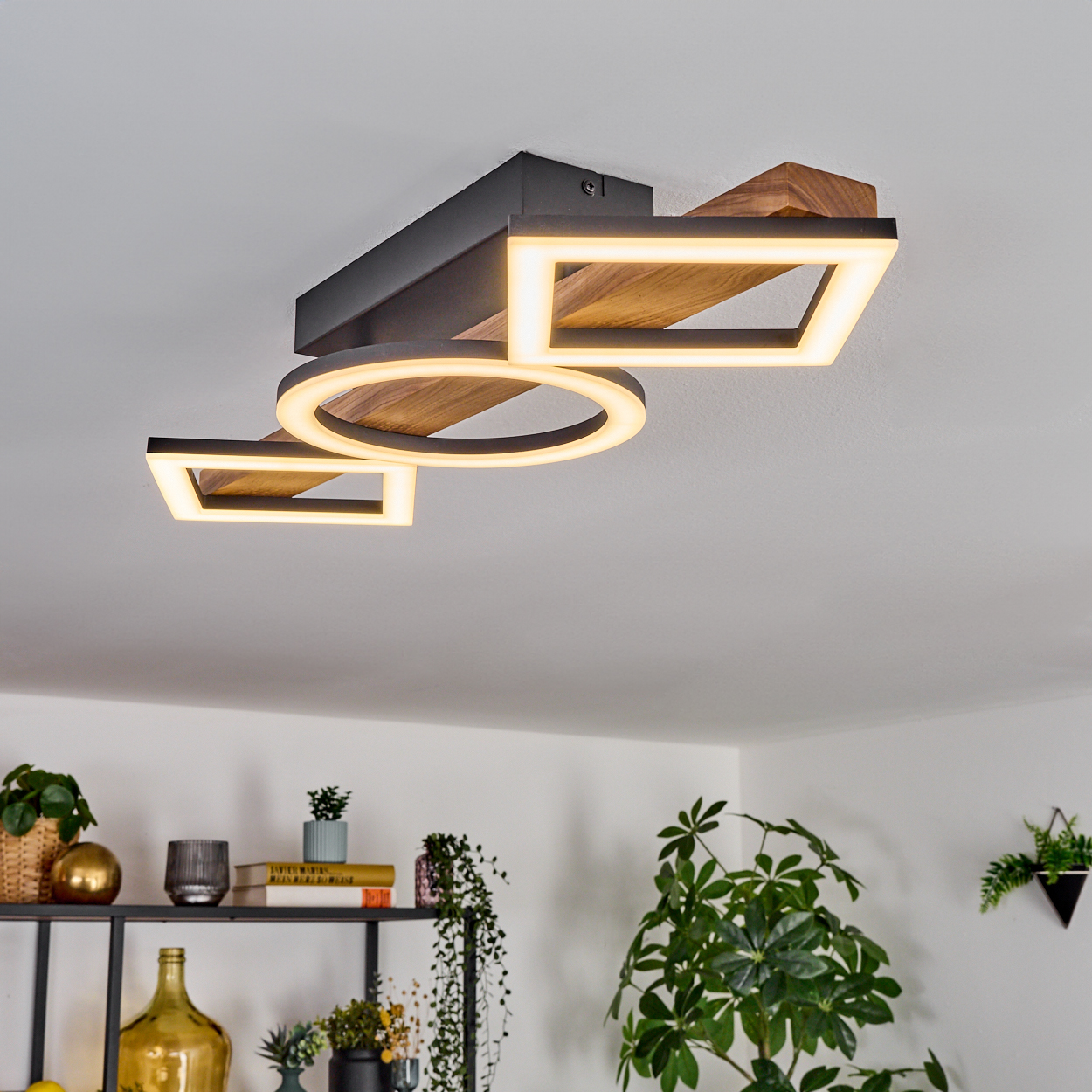 Deckenleuchte mit geometrischen Formen und warmem Licht. Bücherregal mit Pflanzen im Hintergrund.