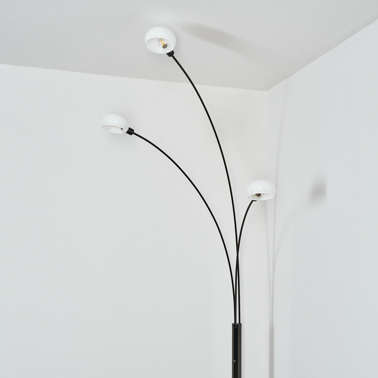 Eine moderne Stehlampe mit drei gebogenen schwarzen Stielen und weißen Lampenköpfen in einer weißen Ecke. Die Wand wirft einen Schatten.