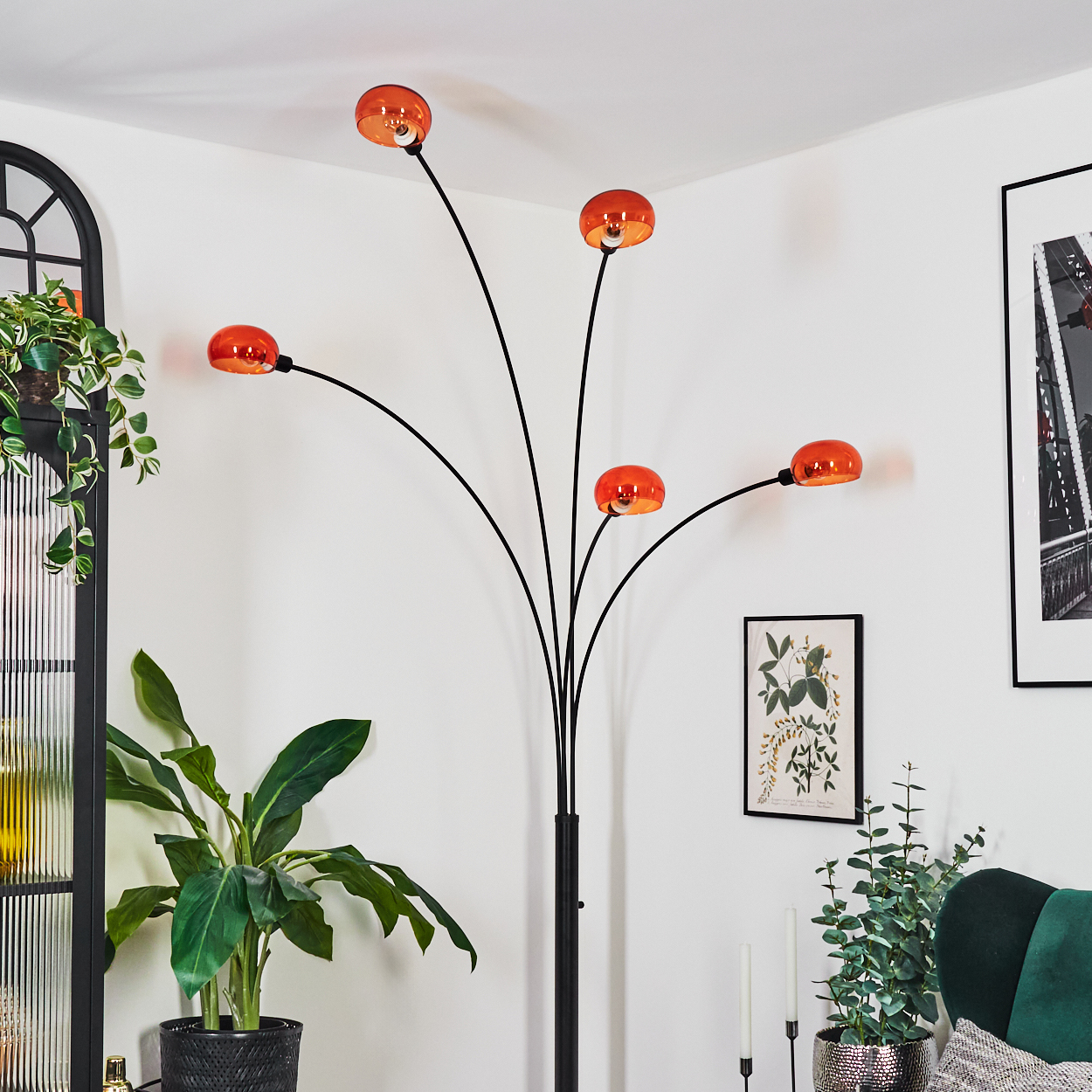 Ein modernes Wohnzimmer mit einer schwarzen Stehlampe mit orangefarbenen Lampenschirmen, Pflanzen und gerahmter Kunst. Weiße Wände und ein grünes Sofa.
