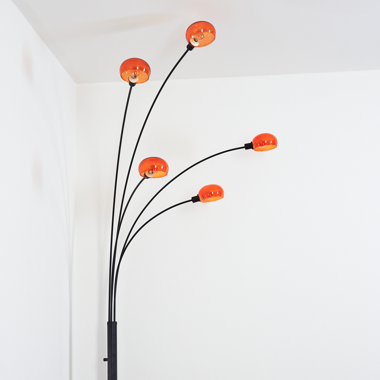 Eine moderne Stehlampe mit fünf orangefarbenen Glasschirmen. Die Lampe hat schwarze Stiele und einen dunklen Sockel, vor einer weißen Wand.