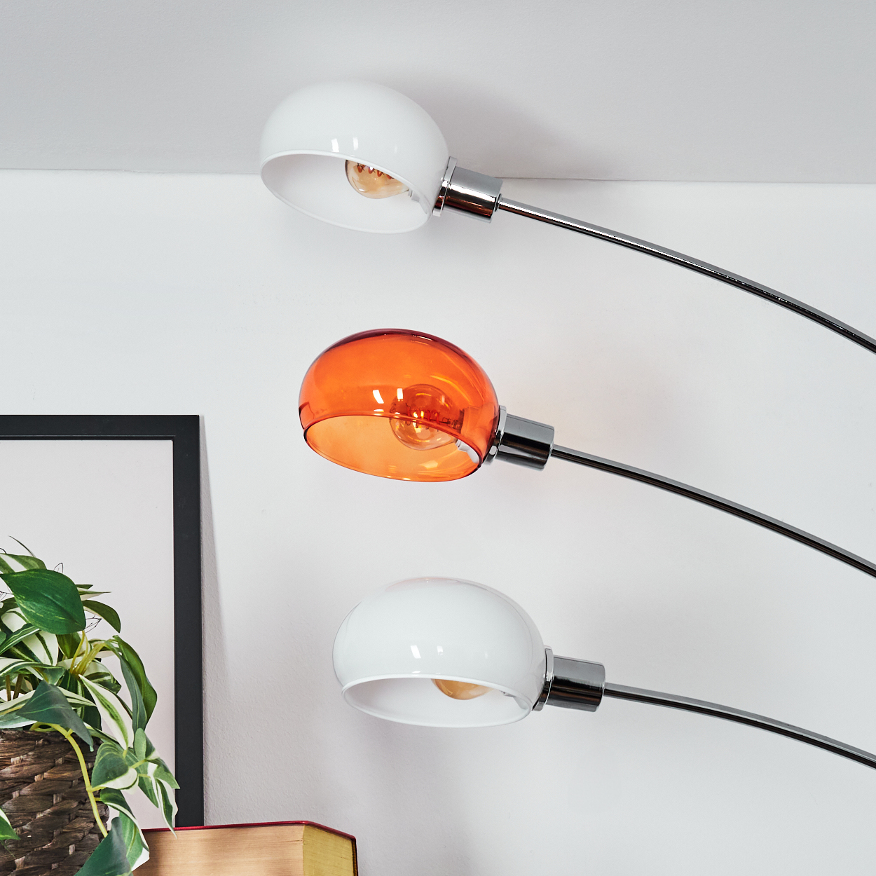 Drei Lampen, zwei weiße und eine orangefarbene, vor einer weißen Wand. Im Hintergrund ein gerahmtes Bild und eine Pflanze.