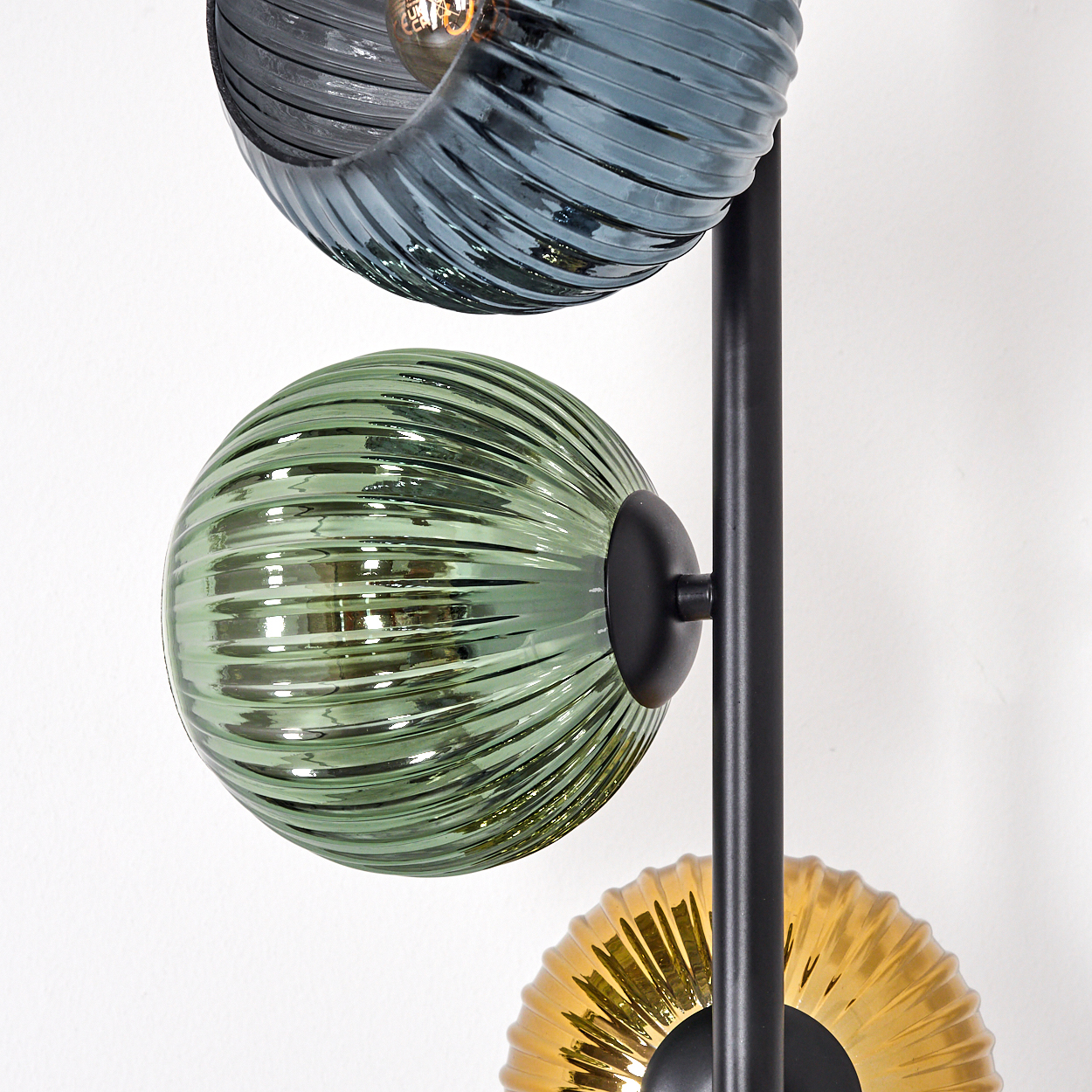 Nahaufnahme einer modernen Lampe mit drei Glaskugeln in Blau, Grün und Gold. Die Lampe hat einen schwarzen Metallrahmen, vor weißer Wand.