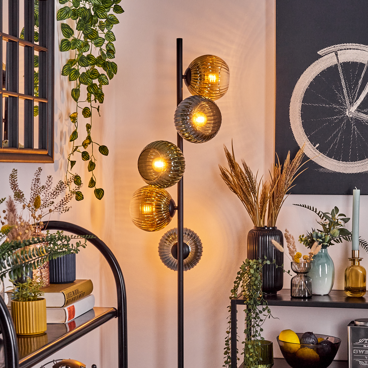 Eine Stehlampe mit Kugelschirmen leuchtet. Daneben befinden sich Pflanzen, ein Regal mit Dekoration und eine Wandkunst mit einem Fahrradrad.