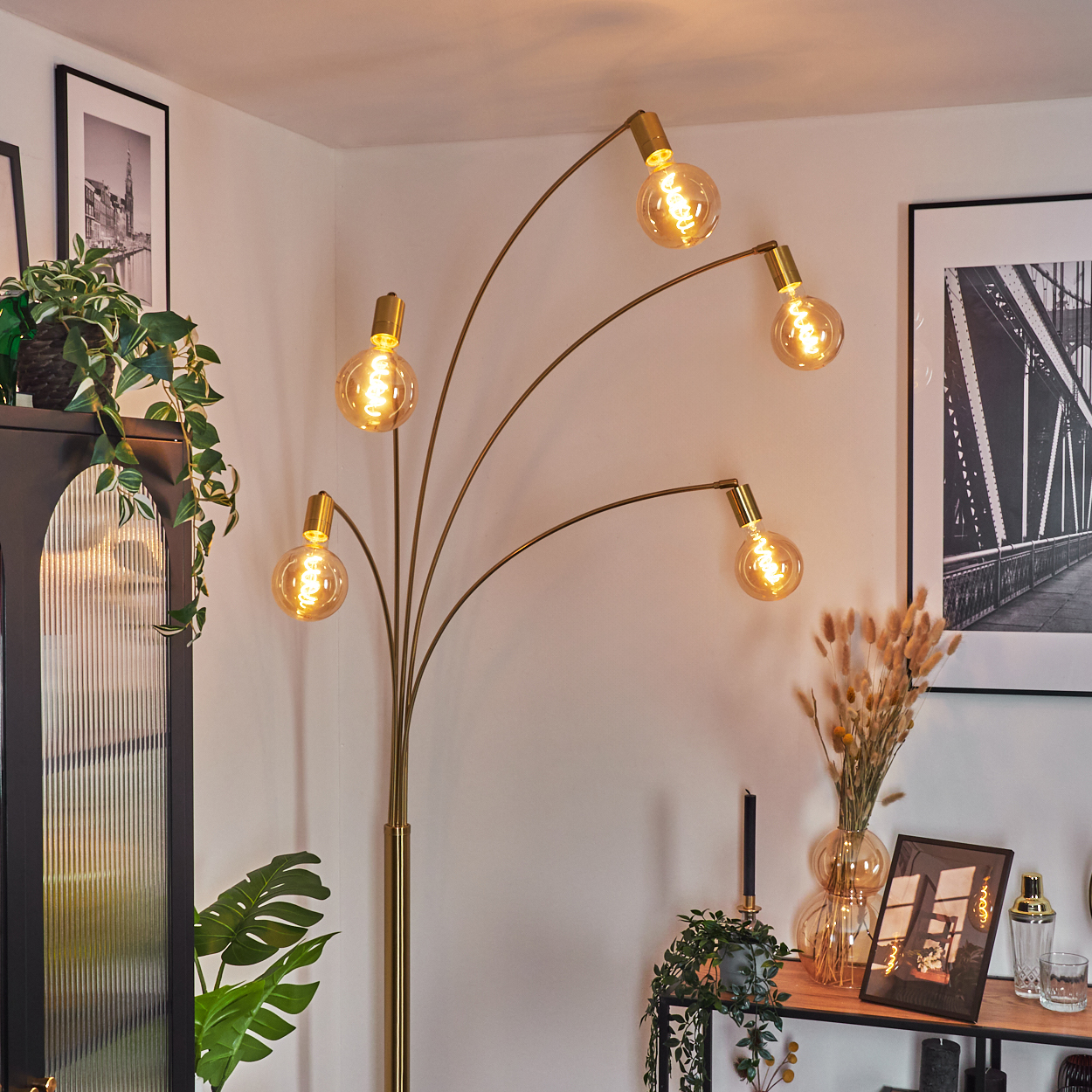 Eine goldene Stehlampe mit fünf Glühbirnen beleuchtet einen Raum. Es gibt einen schwarzen Schrank, Pflanzen, gerahmte Kunst und Dekorationsartikel.