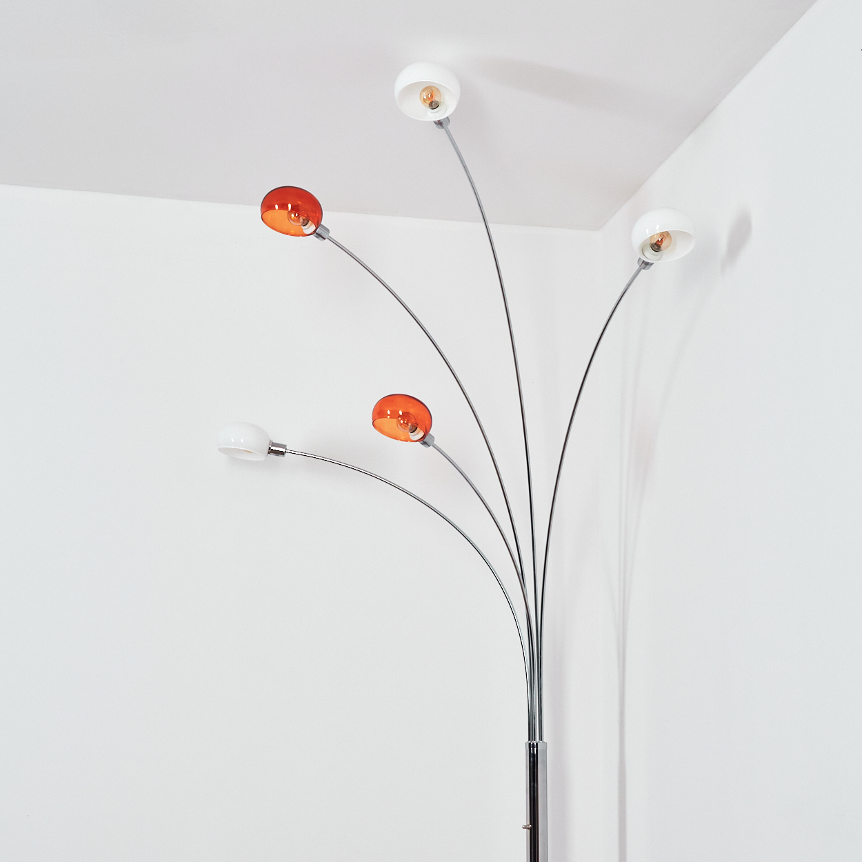 Eine moderne Stehlampe mit fünf gebogenen Armen. Einige Lampen haben orangefarbene Schirme, andere weiße. Sie steht in einem weißen Raum.