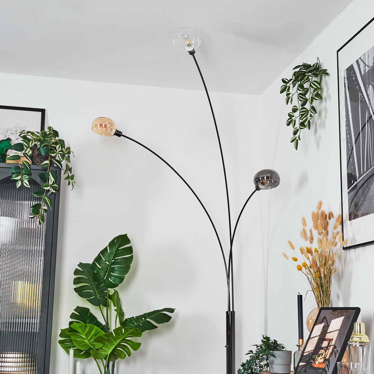 Eine moderne Stehlampe mit drei Glühbirnen, Pflanzen und gerahmten Bildern schmücken den weiß getünchten Raum.