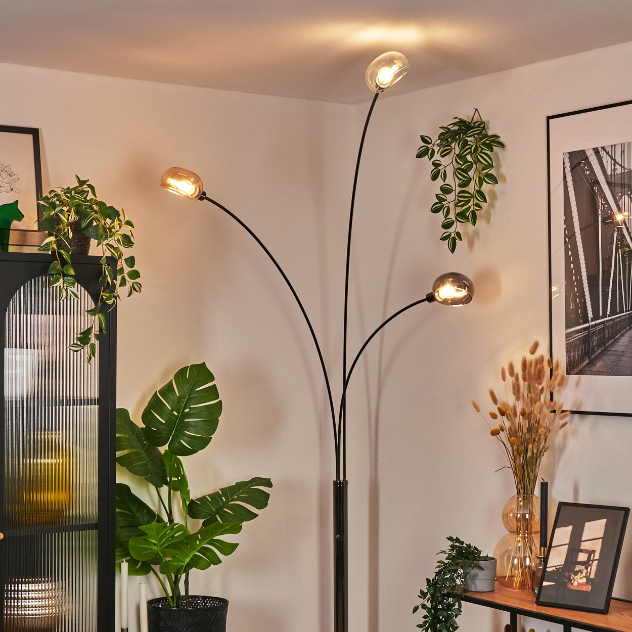 Eine moderne Stehlampe mit drei Glühbirnen, umgeben von Pflanzen und Dekorationen, vor einer weißen Wand.
