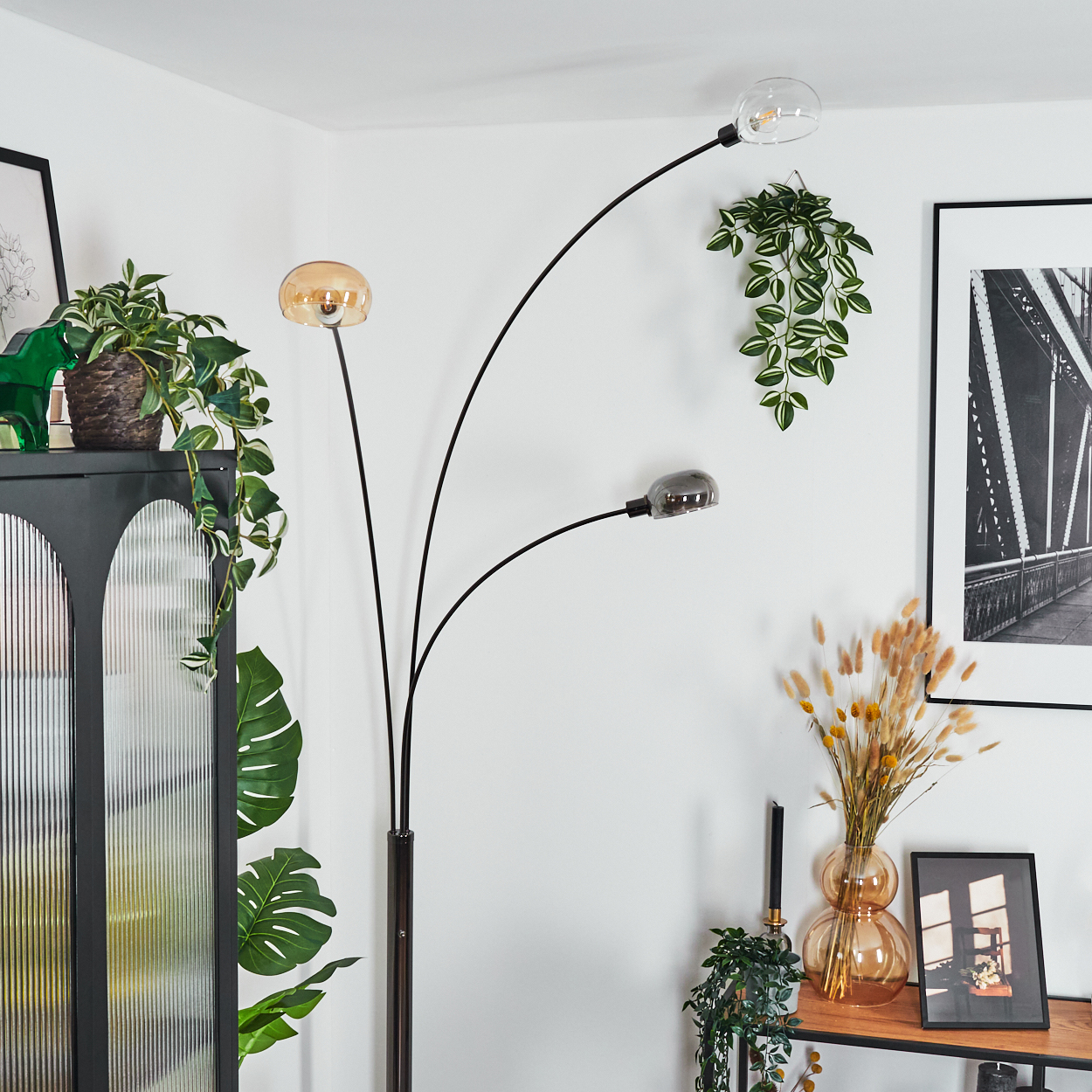 Eine moderne Stehlampe mit drei Lichtern. Pflanzen und Dekorationen schmücken den Raum, darunter ein schwarz-weiß gerahmtes Bild und ein Schrank.