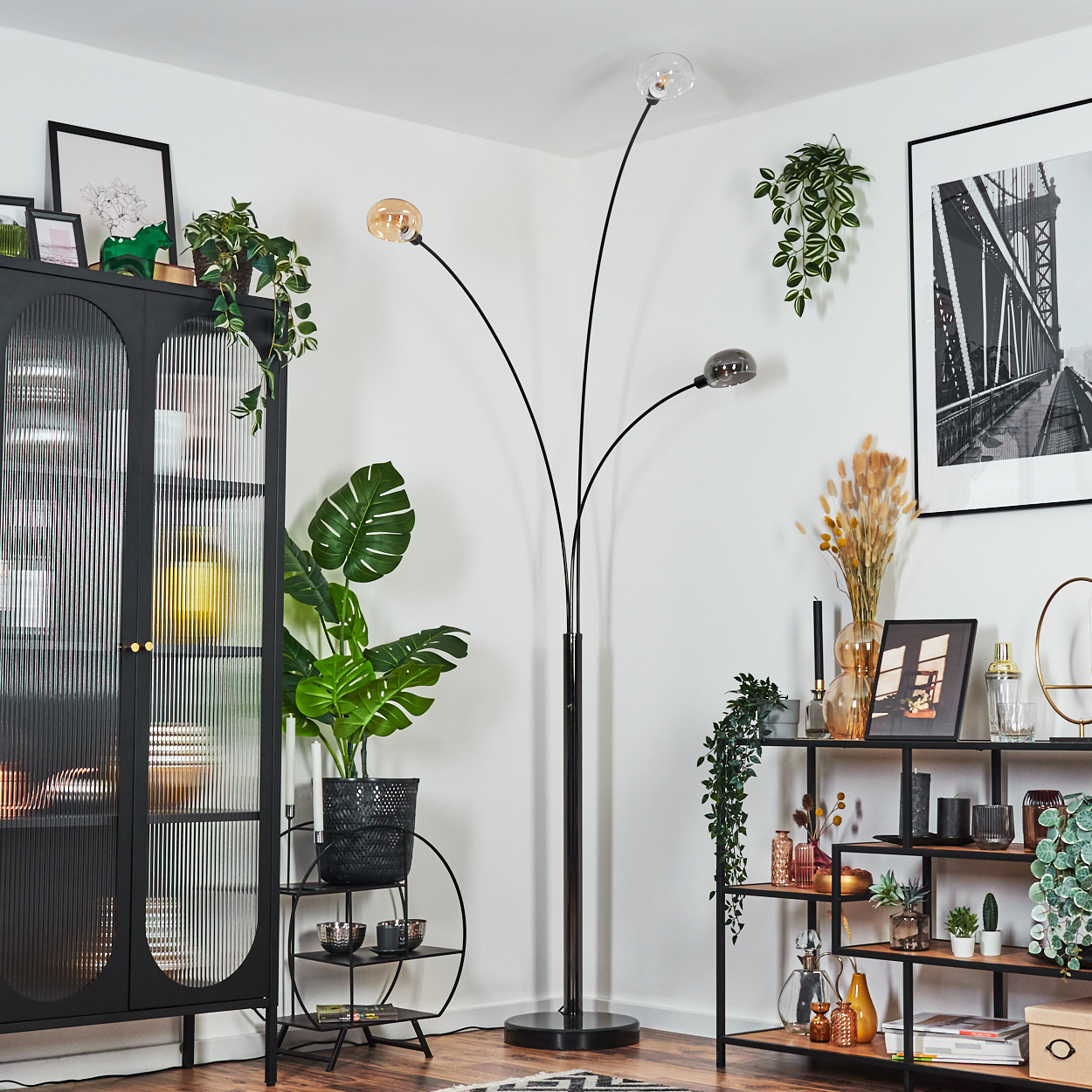 Ein modernes Wohnzimmer mit schwarzem Schrank, Stehlampe und Regalen. Pflanzen und gerahmte Kunst schmücken die weißen Wände.