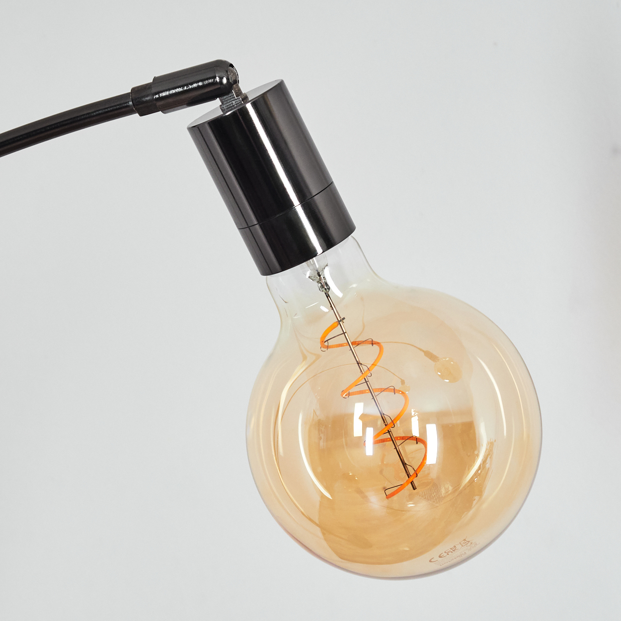 Nahaufnahme einer großen Glühbirne. Sie hat einen orangefarbenen Glühfaden in einem Glaskolben und ist an einem schwarzen Metallarm befestigt.