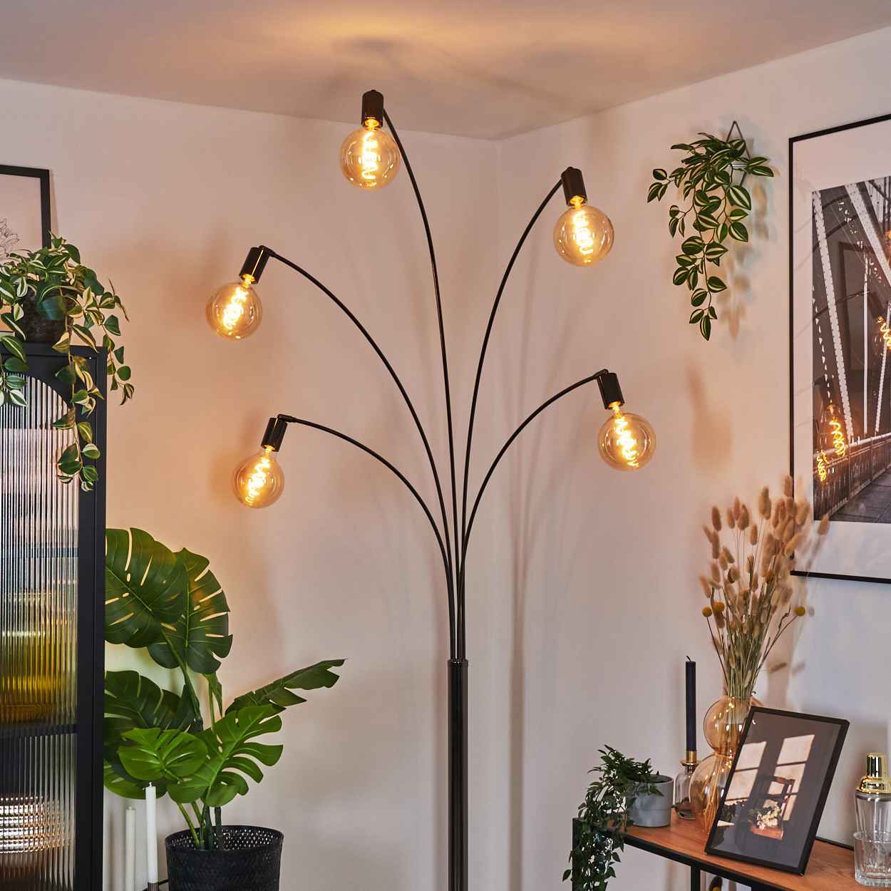 Eine moderne schwarze Stehlampe mit fünf freiliegenden Glühbirnen. Der Raum hat Pflanzen, ein Bild und ein Regal.