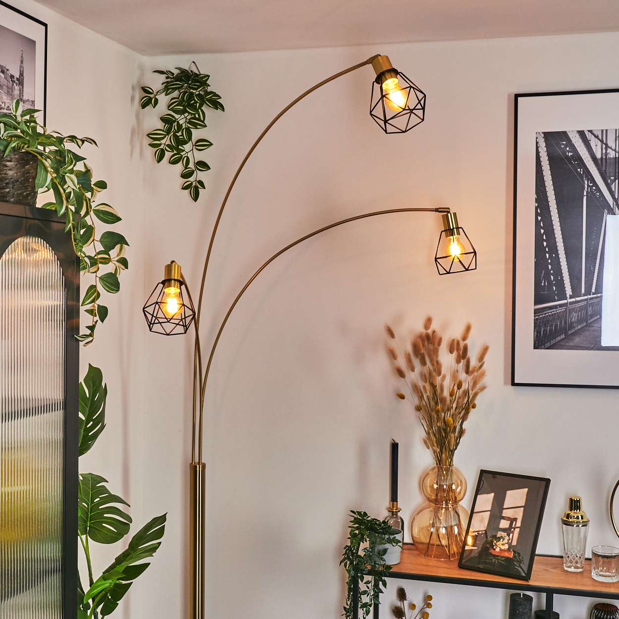 Goldene Stehlampe mit drei Lichtern. Pflanzen und Bilder schmücken den Raum. Auf der rechten Seite eine schwarze Kerze.