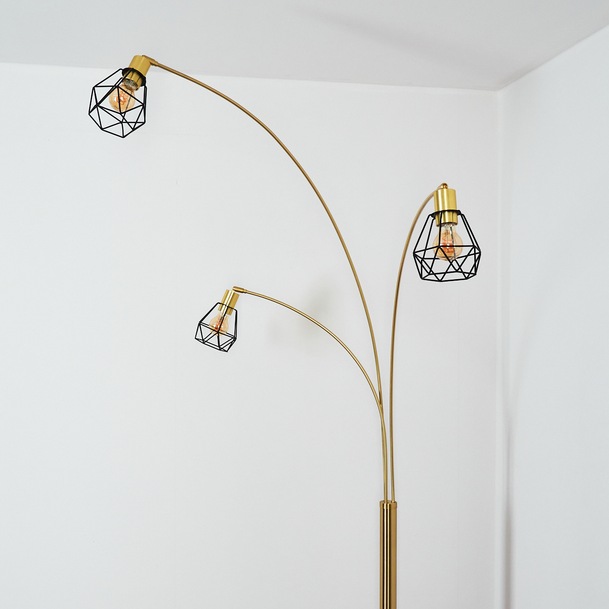 Eine goldene Stehlampe mit drei schwarzen Lampenschirmen im Käfigstil. Die Glühbirnen sind beleuchtet, der Hintergrund ist eine weiße Wand.