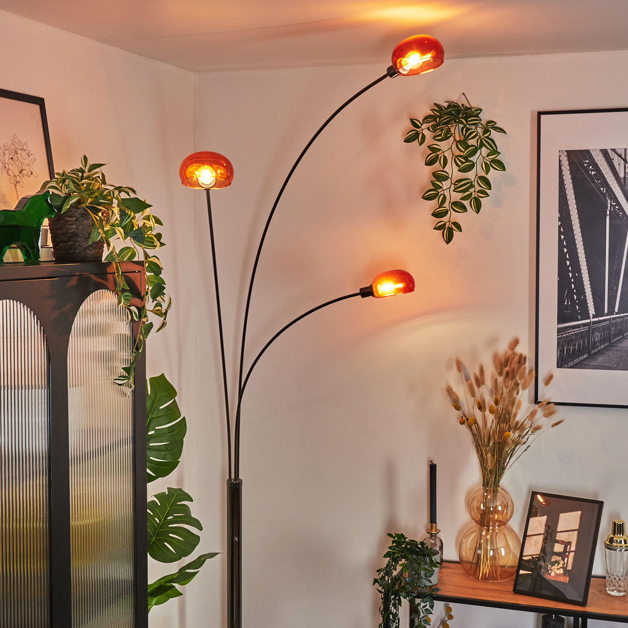 Ein Raum mit einer Stehlampe mit drei orangefarbenen Lampenschirmen. Pflanzen und Dekorationsartikel sind um die Lampe angeordnet.
