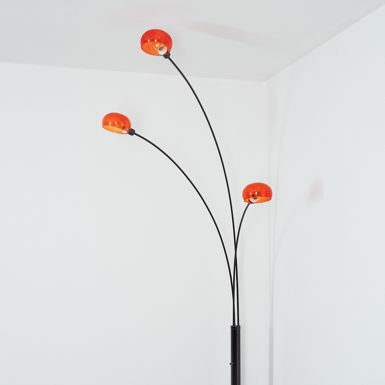 Eine moderne Stehlampe mit drei gebogenen schwarzen Armen. Jeder Arm hat einen runden, orangefarbenen Lampenschirm. Der Hintergrund ist eine weiße Wand.