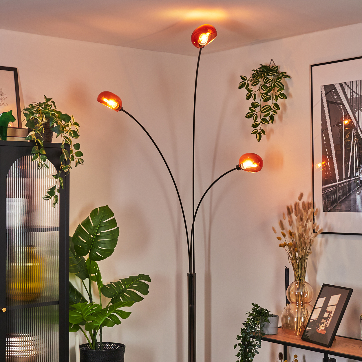 Eine moderne Stehlampe mit vier orangefarbenen Glasschirmen. Pflanzen und Dekorationen sind im Hintergrund, darunter ein schwarzer Schrank und ein gerahmtes Bild.