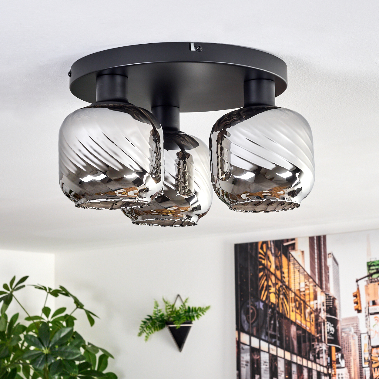 Deckenleuchte mit drei silbernen Glasschirmen. Die Schirme haben ein Spiraldesign. Die Leuchte ist schwarz. Pflanzen und ein Stadtbild sind im Hintergrund.