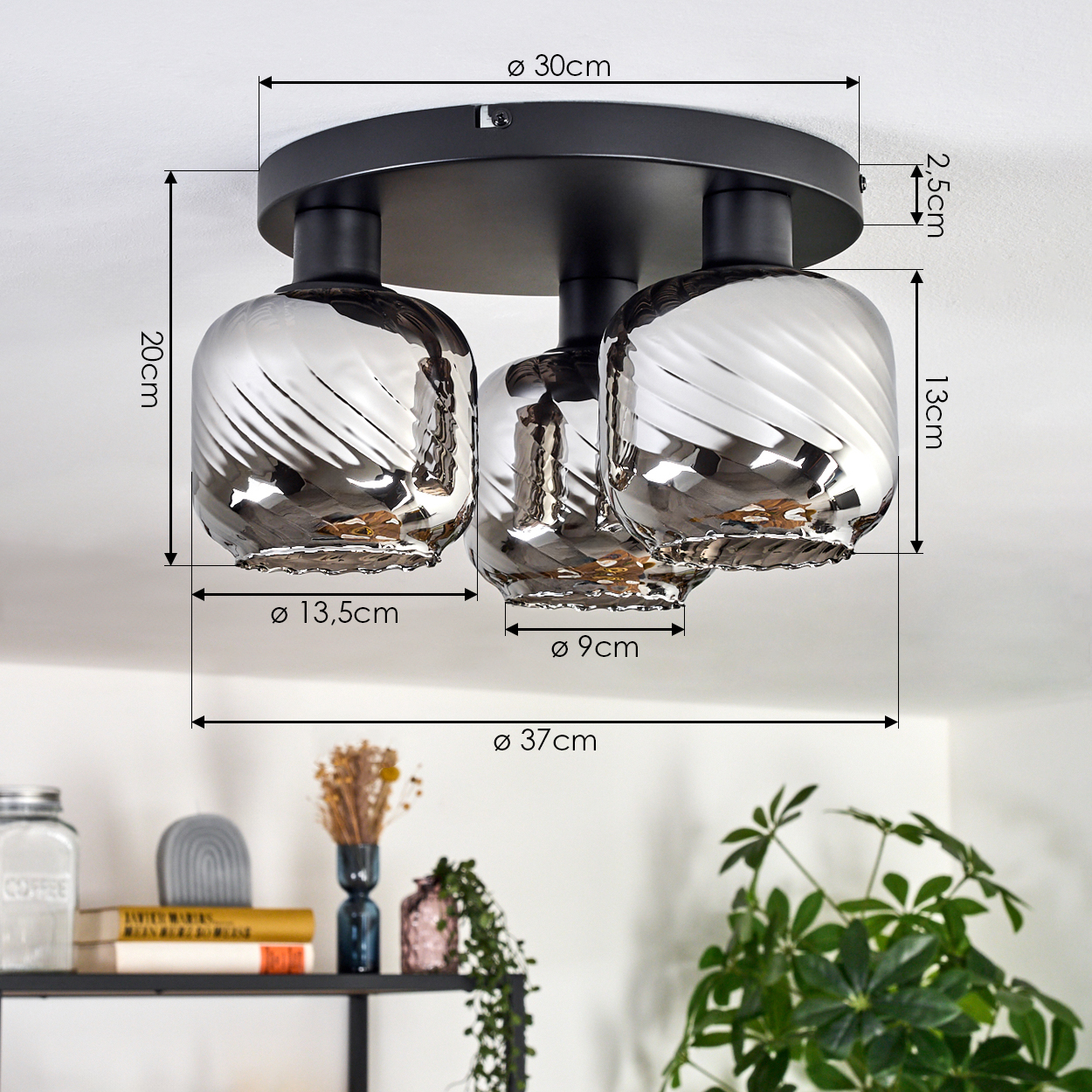 Eine moderne Deckenlampe mit drei silberfarbenen Glasschirmen wird gezeigt. Abmessungen sind beschriftet. Ein Regal mit Dekoration ist im Hintergrund.