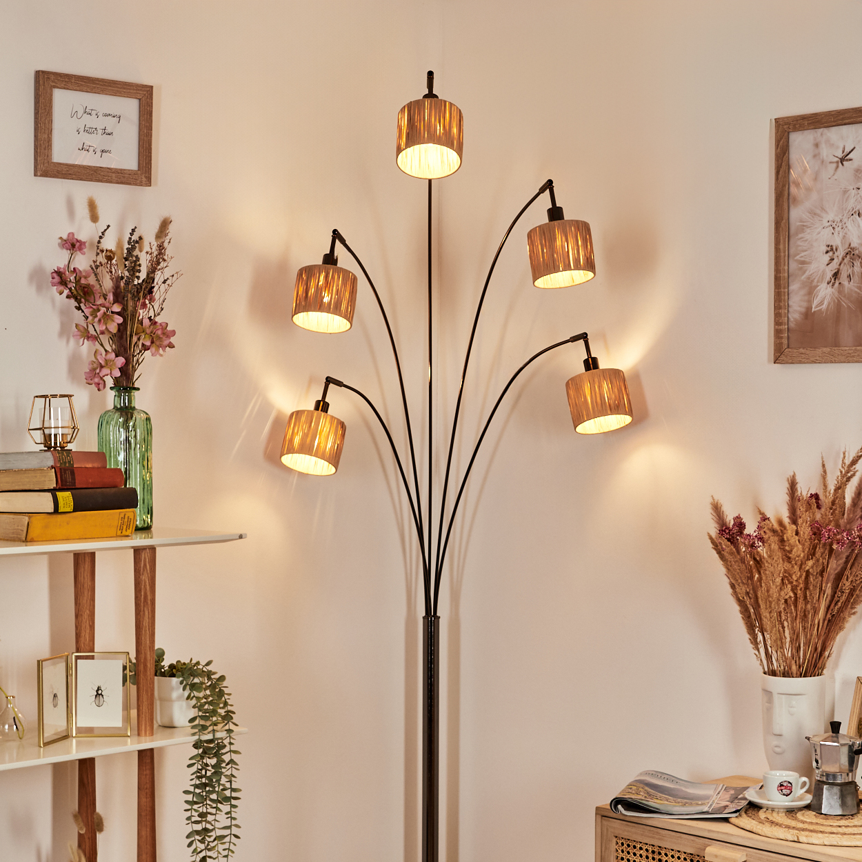 Eine Stehlampe mit fünf Schirmen beleuchtet einen Raum. Regale mit Dekoration befinden sich links. Auf einem Tisch liegen eine Zeitschrift, Kaffee und eine dekorative Vase.