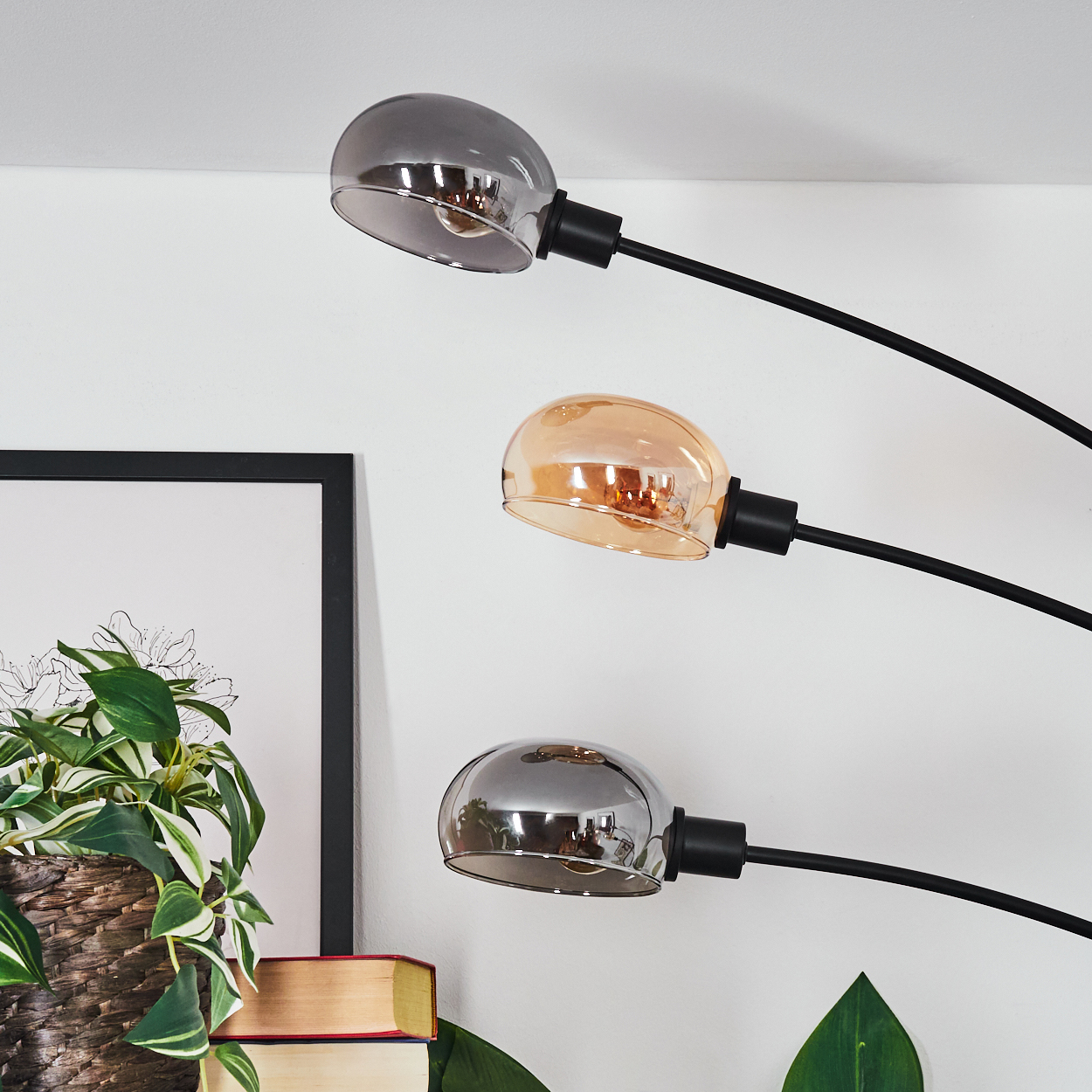 Drei Lampen mit Glasschirmen, schwarze Arme. Eine Pflanze im Korb, Bücher und ein gerahmtes Bild sind im Bild.