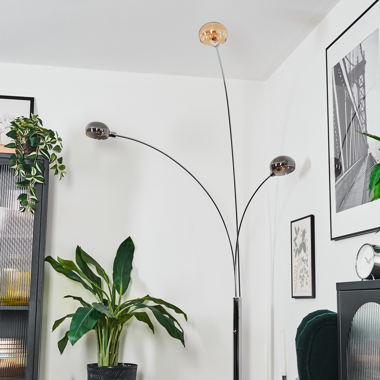 Eine moderne Stehlampe mit drei kugelförmigen Schirmen. Eine große grüne Pflanze in einem Topf steht davor. Im Hintergrund ein schwarzer Schrank und ein gerahmtes Bild.