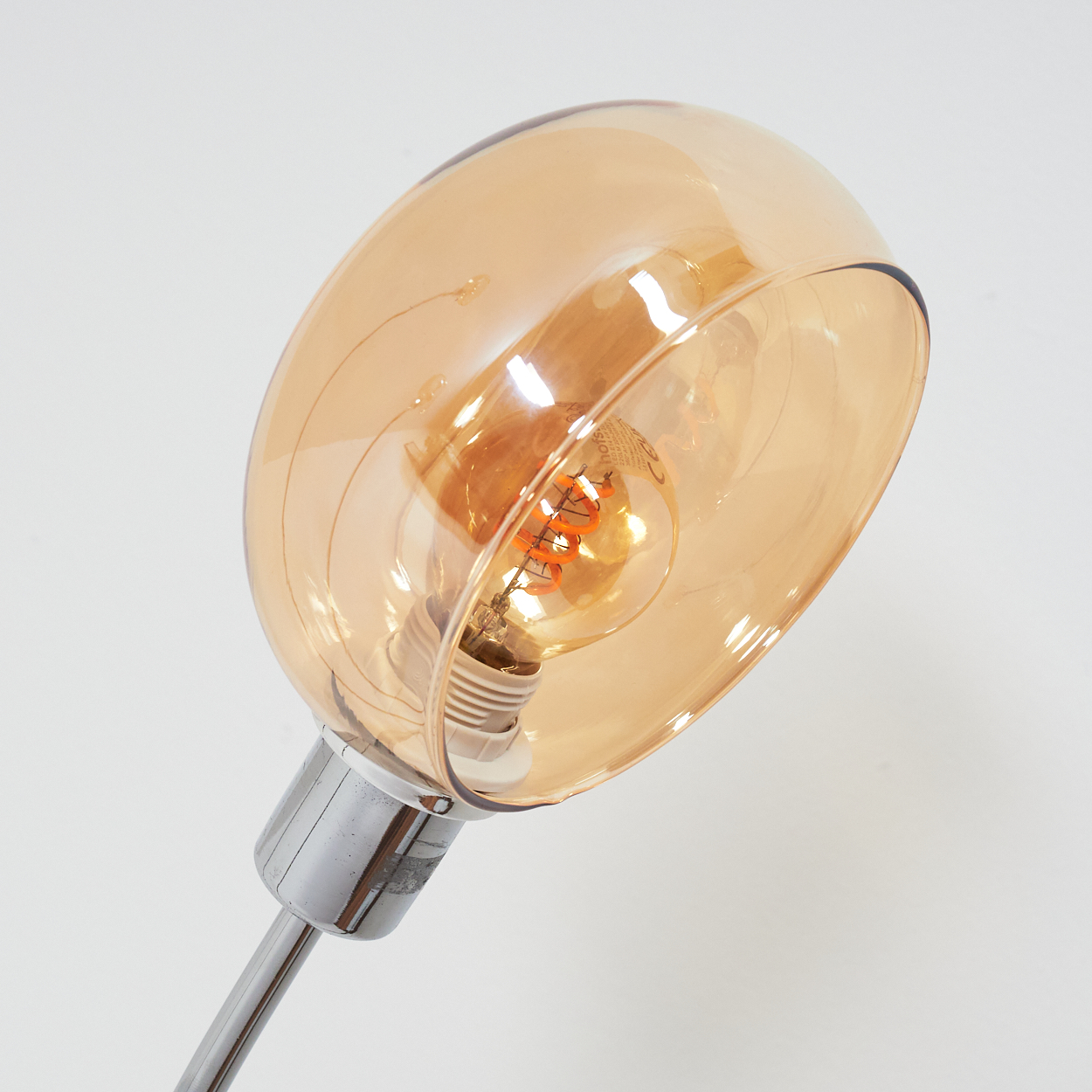 Nahaufnahme einer Lampe mit Chromfuß und einer Glühbirne, die von einem durchscheinenden, bernsteinfarbenen Glasschirm umgeben ist.