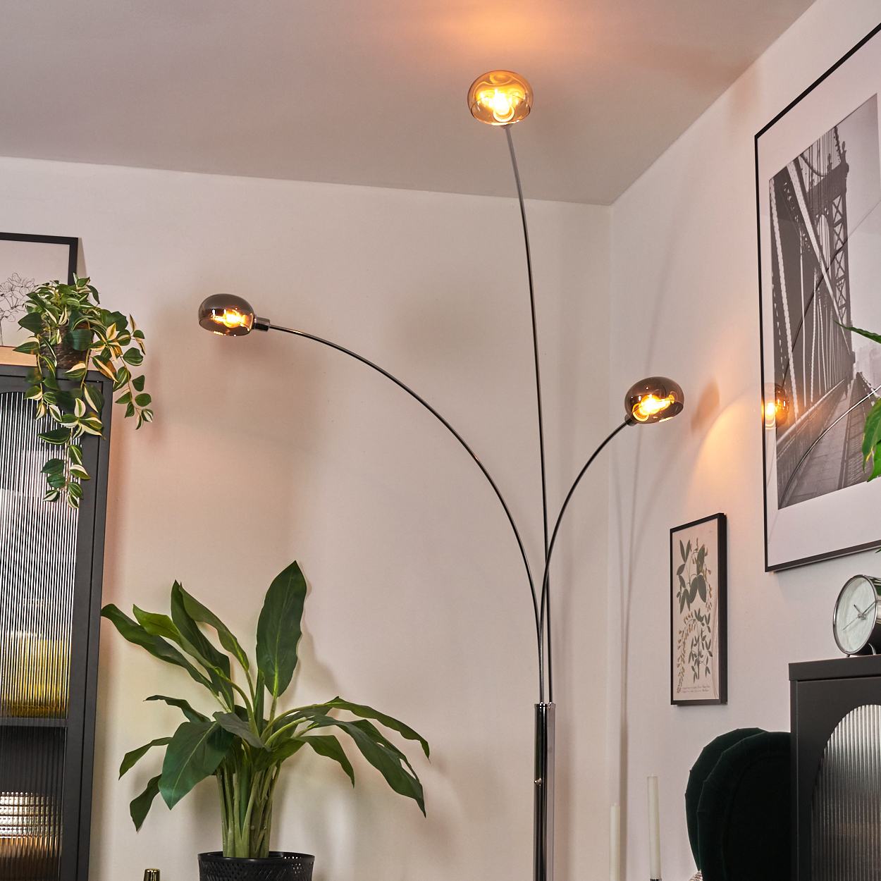Eine moderne Stehlampe mit drei Lichtern. Vor der Lampe steht eine Pflanze. Weitere Pflanzen, ein gerahmtes Bild und eine Uhr sind sichtbar.