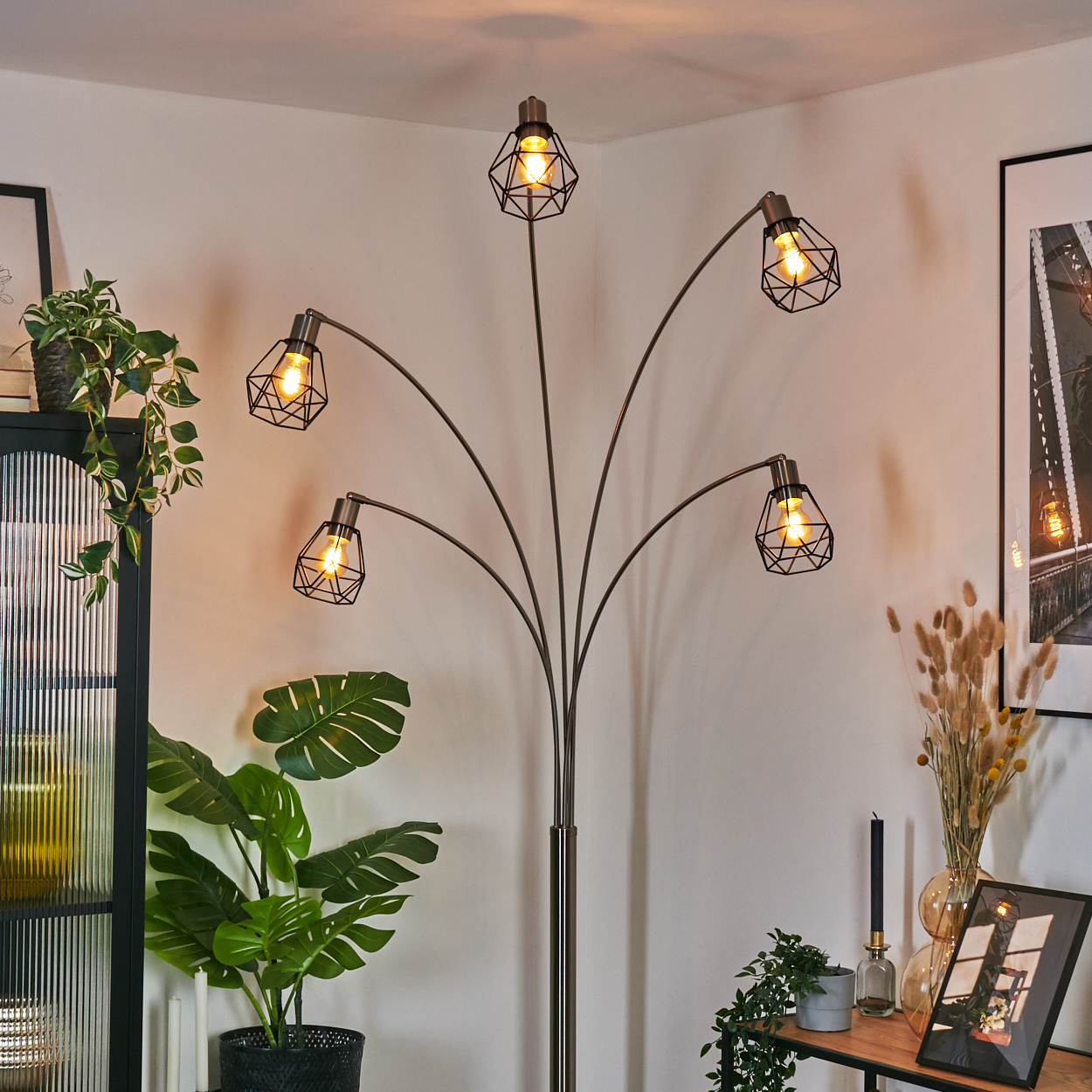 Eine moderne Stehlampe mit fünf Lichtern. Im Hintergrund eine große Pflanze und ein Glasschrank, rechts ein Bilderrahmen.