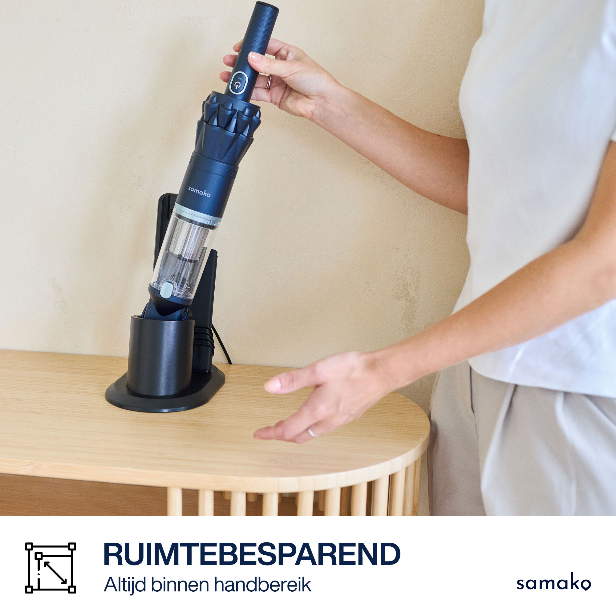 A woman holds a black Samako vacuum. It is on a black stand on a wooden table. Beige wall in the background.