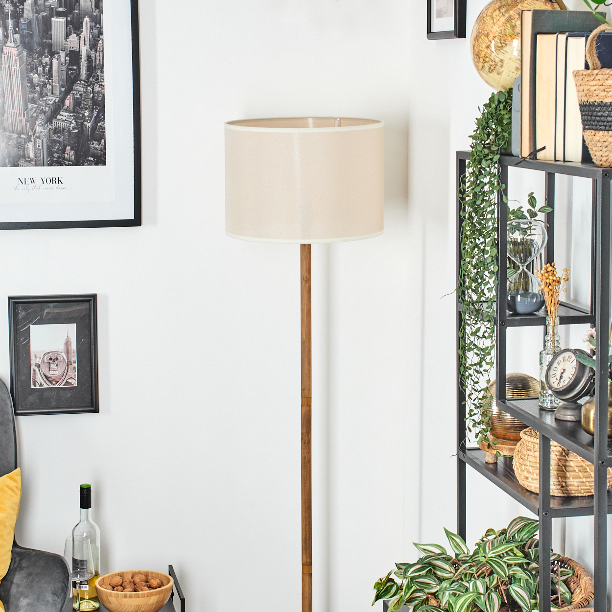 Beiger Lampenschirm auf Holzständer. Regal mit Büchern, Pflanzen und Dekorationen. New York-Poster und gerahmtes Foto an der Wand.